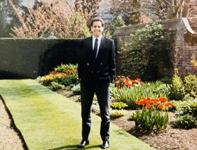 Jonathan stood in a suit in a beautiful garden smiling. There are orange and yellow flowers surrounding the border.