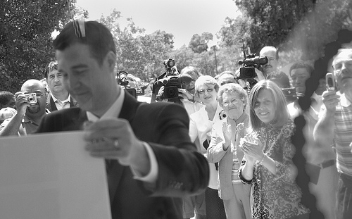 Black and white photo of Dayne stood before a crowd putting a sign up (unpictured). The crowd behind him are smiling, clapping, and taking photographs.