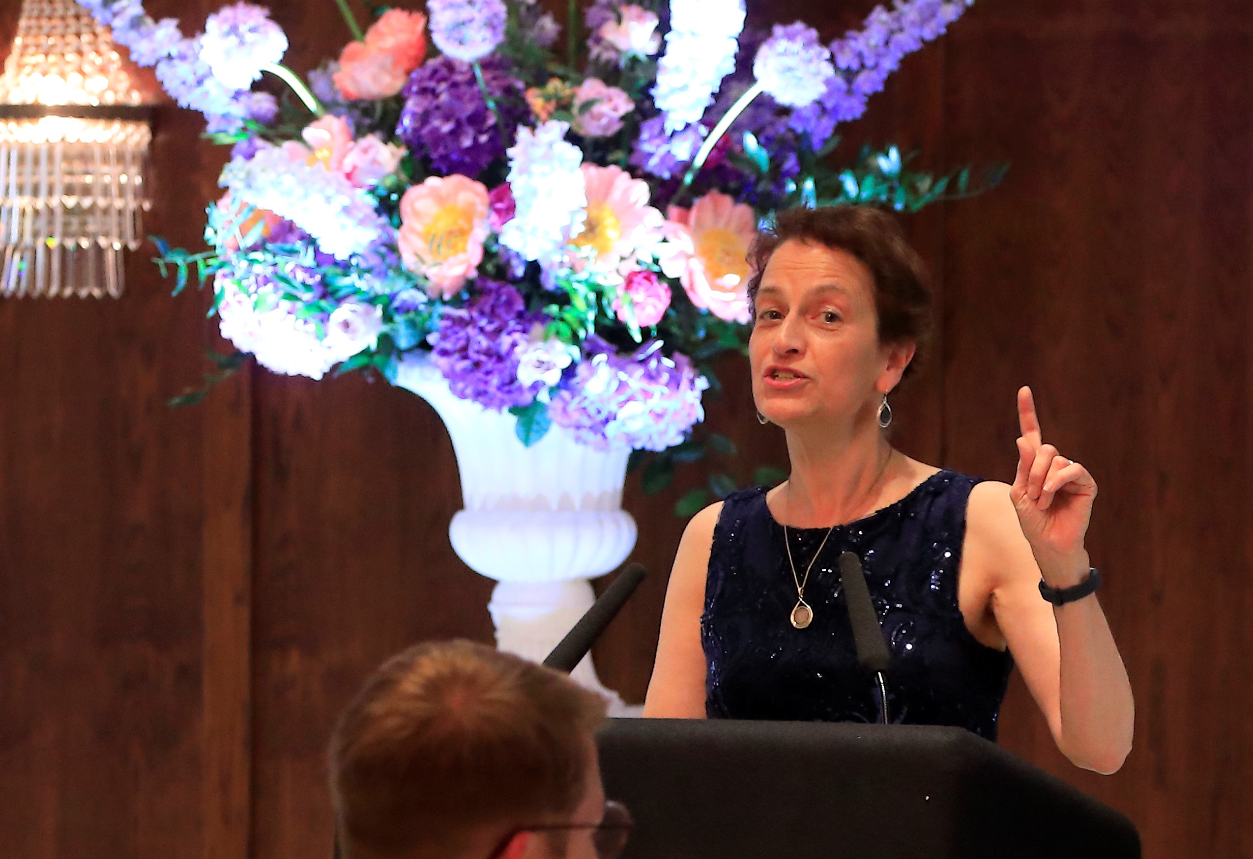Image of a woman stood at a podium in a dark blue dress making a speech to seated attendees. There is a bouquet of flowers behind her. 