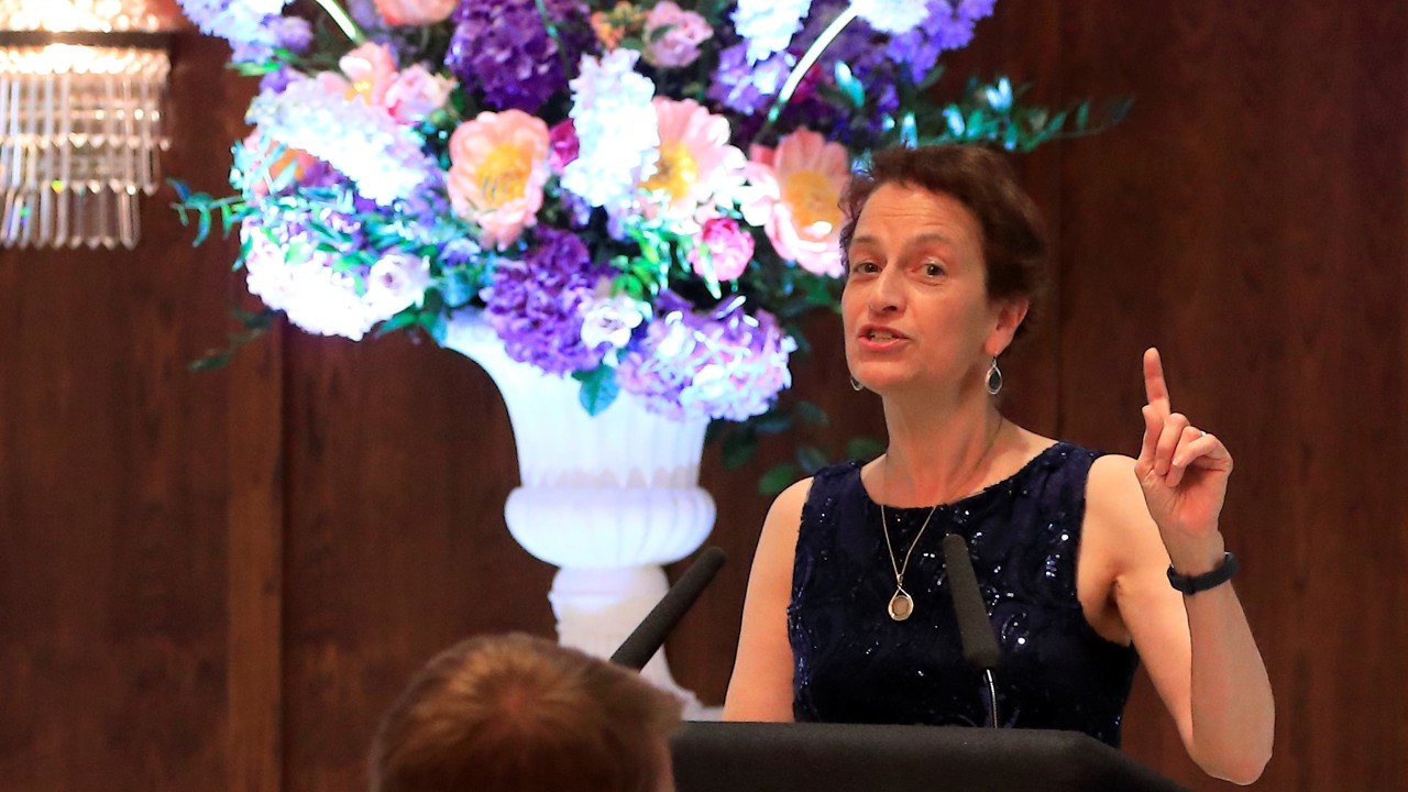 Image of a woman stood at a podium in a dark blue dress making a speech to seated attendees. There is a bouquet of flowers behind her.