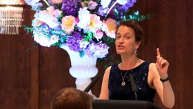 Image of a woman stood at a podium in a dark blue dress making a speech to seated attendees. There is a bouquet of flowers behind her.