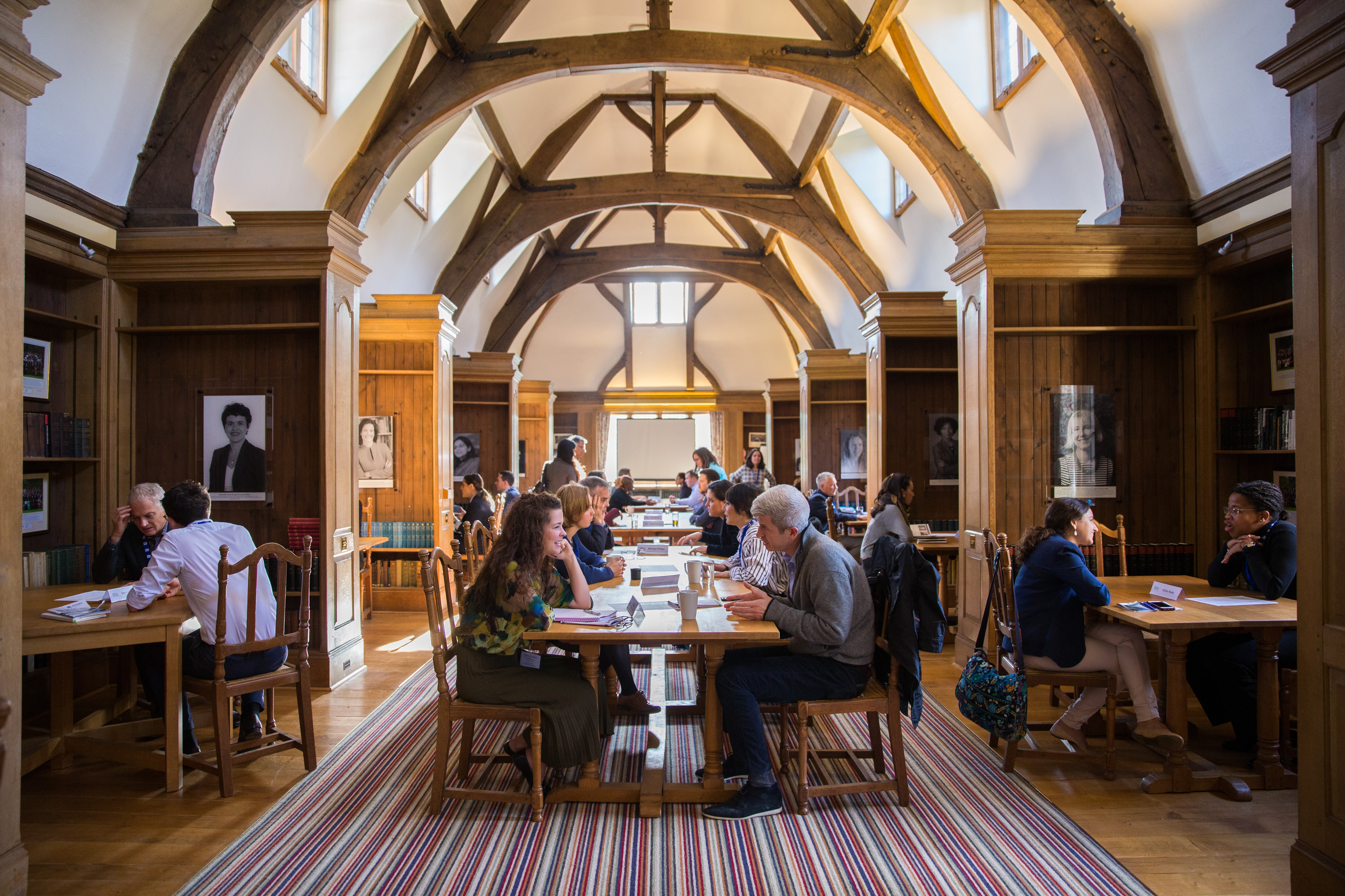 A wide shot of several people sitting at tables in a library. Most are sat in pairs and are in the middle of a discussion.