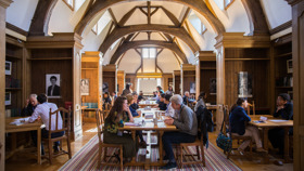 A wide shot of several people sitting at tables in a library. Most are sat in pairs and are in the middle of a discussion.