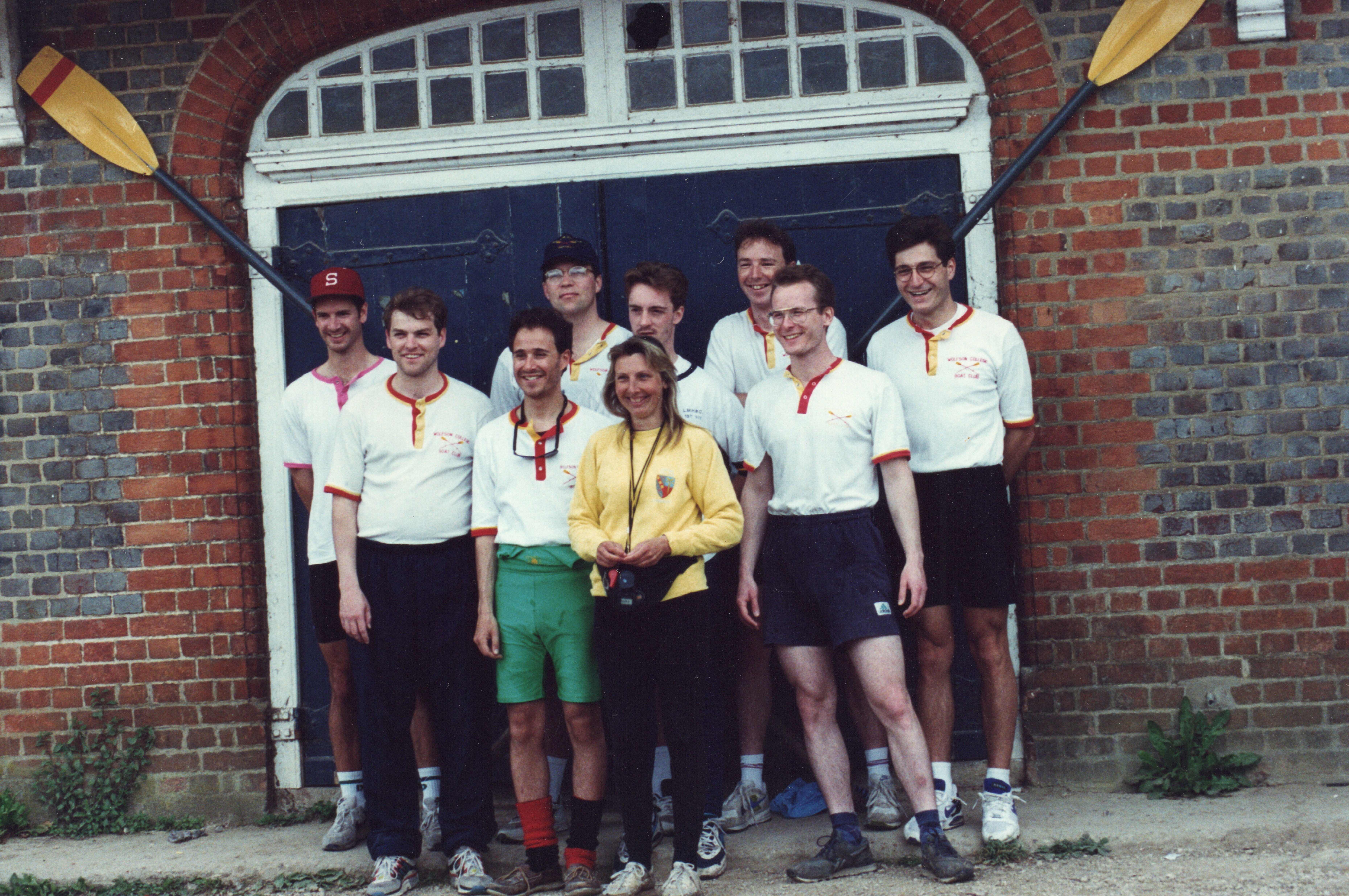 A group of 9 rowers posing for a group photo, all wearing their uniform which is a white polo shirt and shorts.