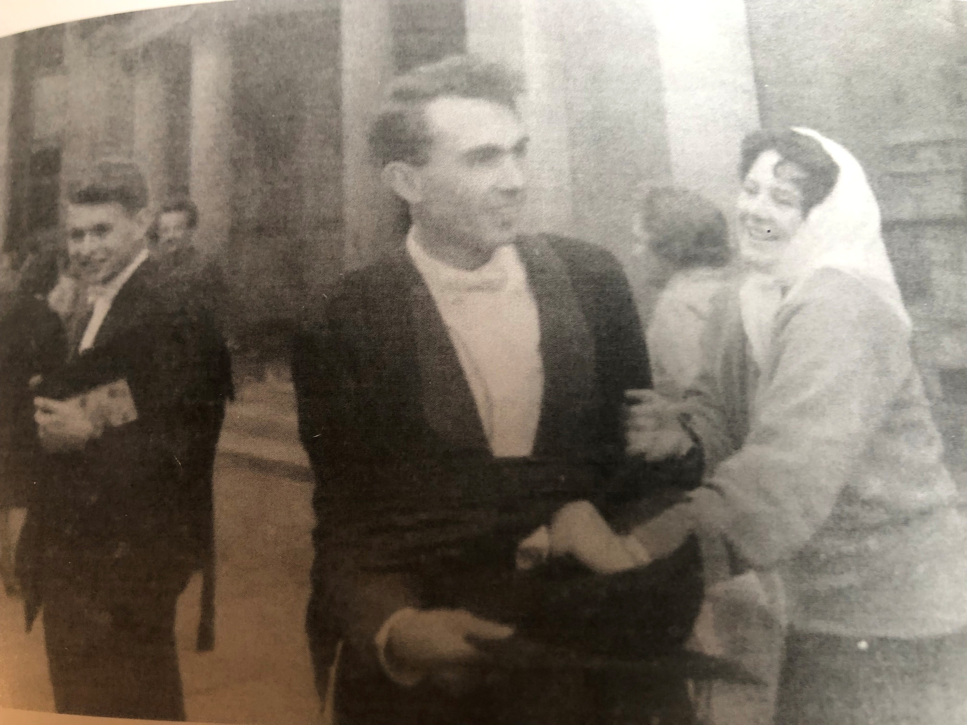 Black and white photo of John wearing his Oxford gown. He is being grabbed by a smiling woman wearing a white head scarf.