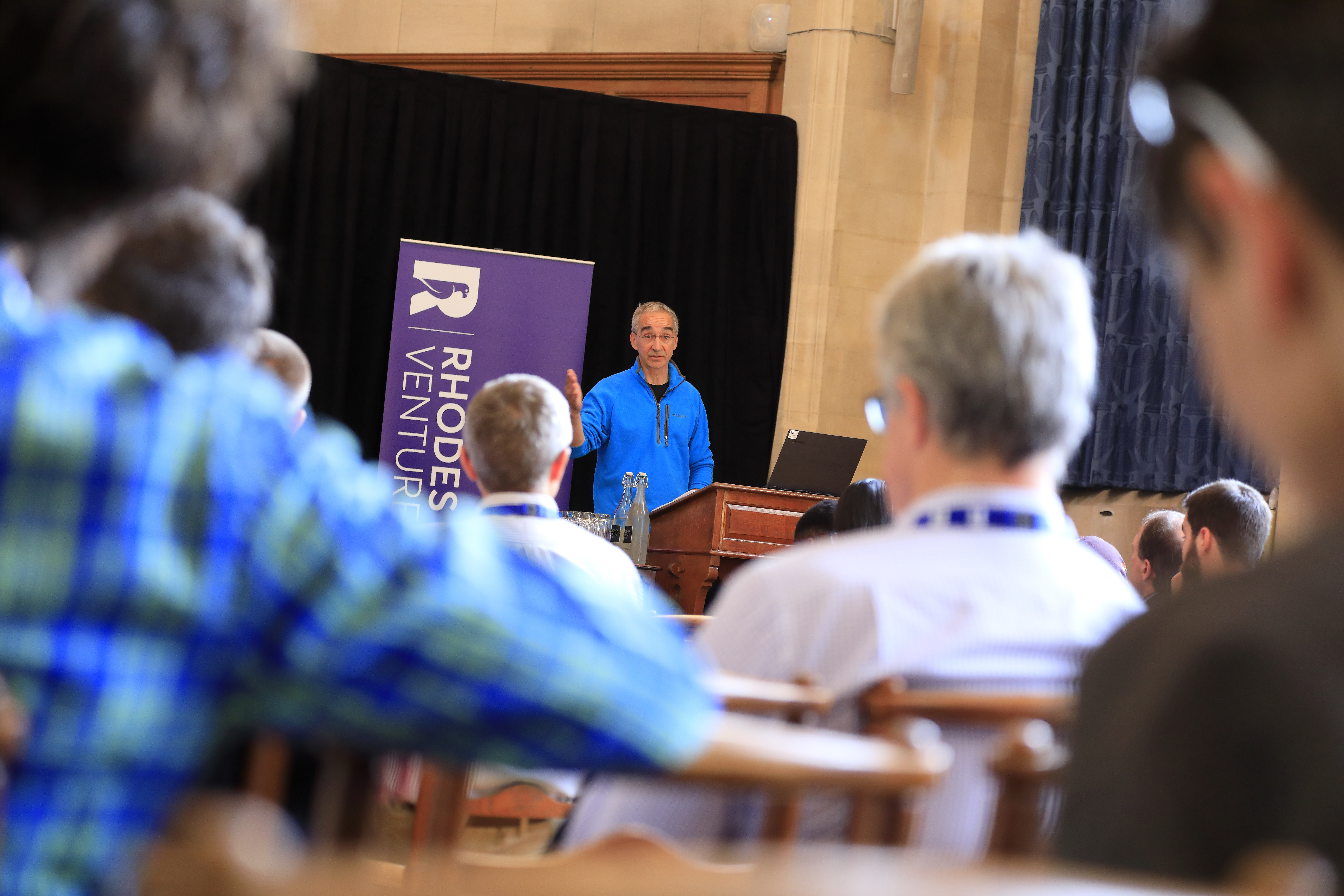 Image of a man in a blue jumper standing at the front of a lecture hall speaking to a room of seated attendees. A sign saying Rhodes Ventures is in the background. 