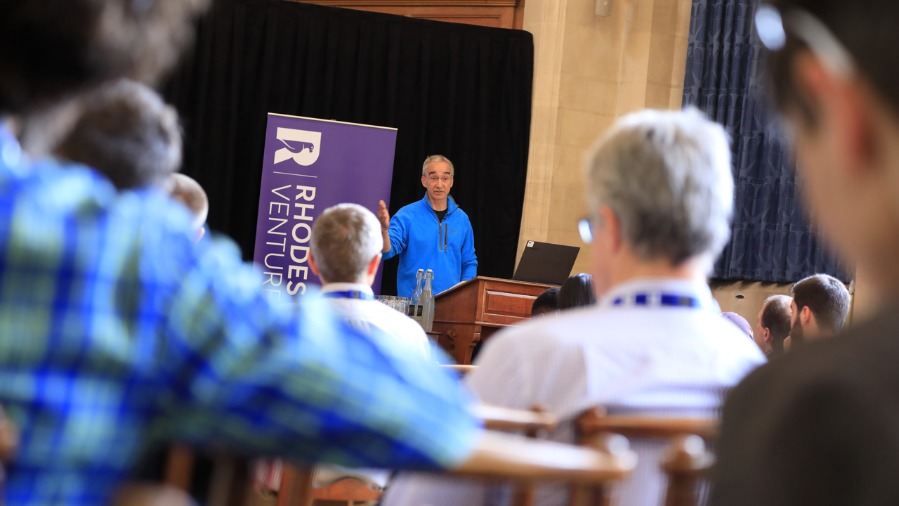 Image of a man in a blue jumper standing at the front of a lecture hall speaking to a room of seated attendees. A sign saying Rhodes Ventures is in the background.