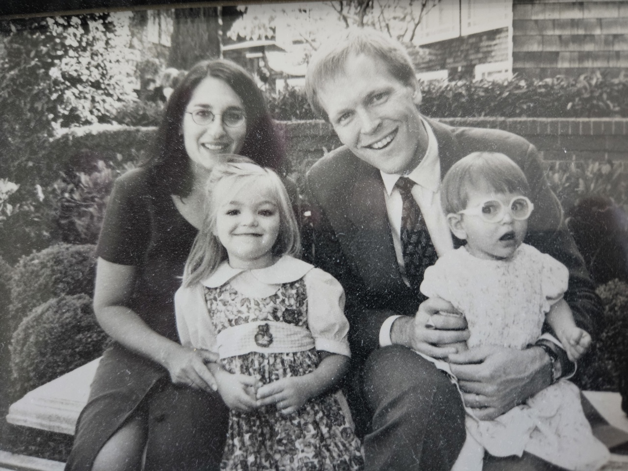 Black and white family photo taken on a bench. A mother and a father sit and smile on the bench holding their younger children, one girl and one boy.