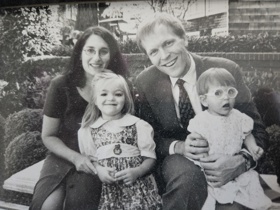 Black and white family photo taken on a bench. A mother and a father sit and smile on the bench holding their younger children, one girl and one boy.