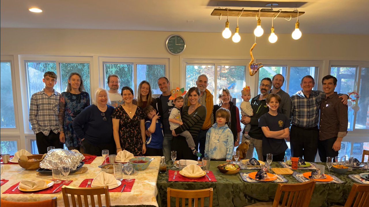 The picture shows a larger family standing behind a table. There are plates, cutlery and food on the table.