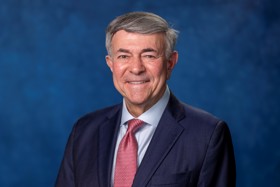 Bob wearing a black suit, smiling into camera, in front of a blue background.