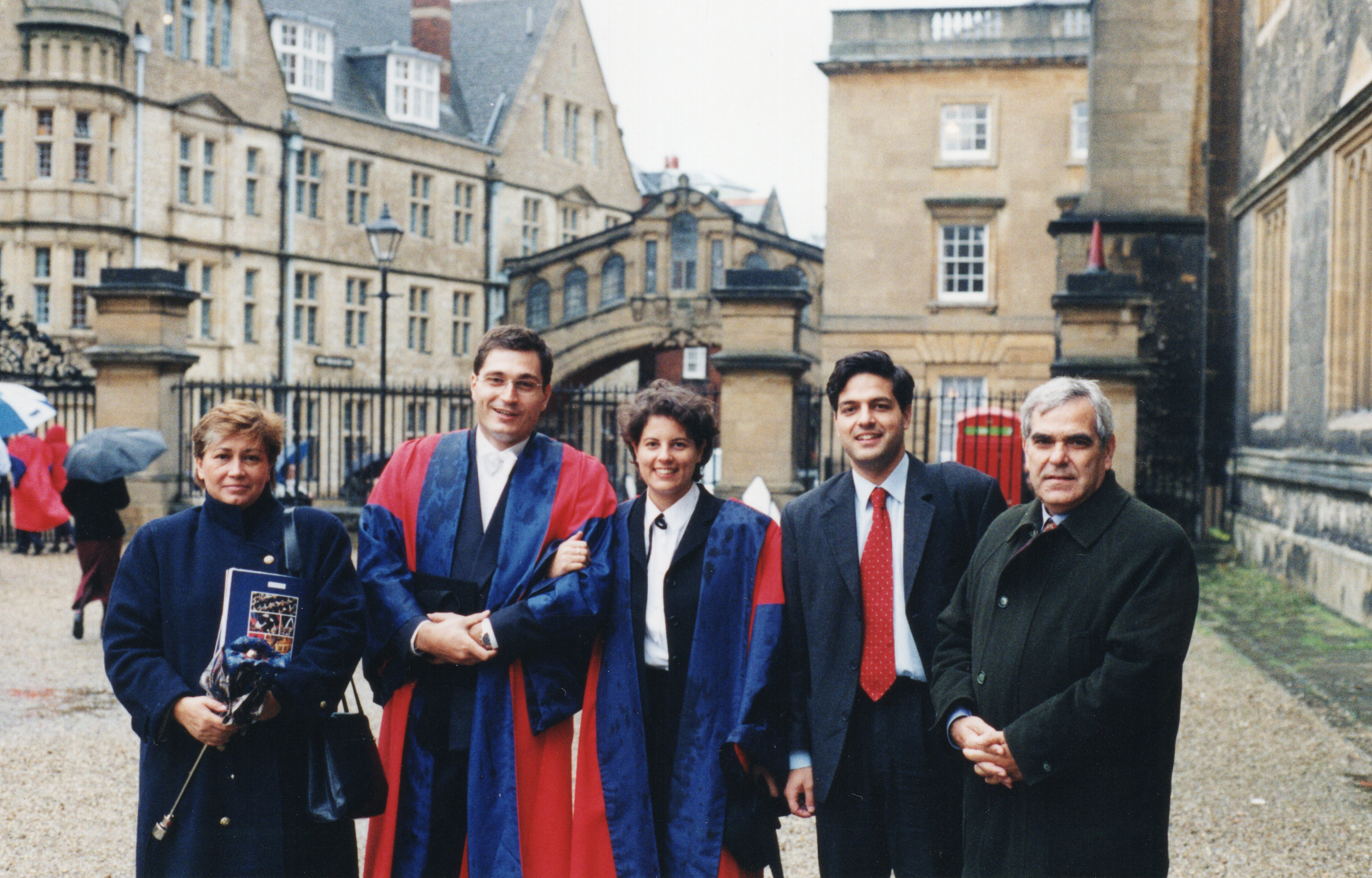 Group of 5 stood before the Oxford Bridge of Sighs. Argyris is second to the left. Him and a young lady are both wearing blue and red matriculation gowns, and the rest of the group is in formal attire.