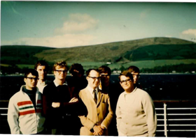 The picture shows a group of people in warm clothing, aboard a ship. There are the sea and the hills in the background.