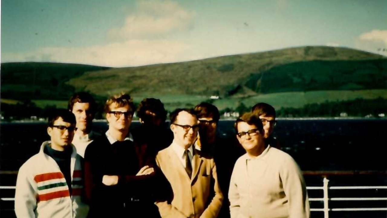 The picture shows a group of people in warm clothing, aboard a ship. There are the sea and the hills in the background.