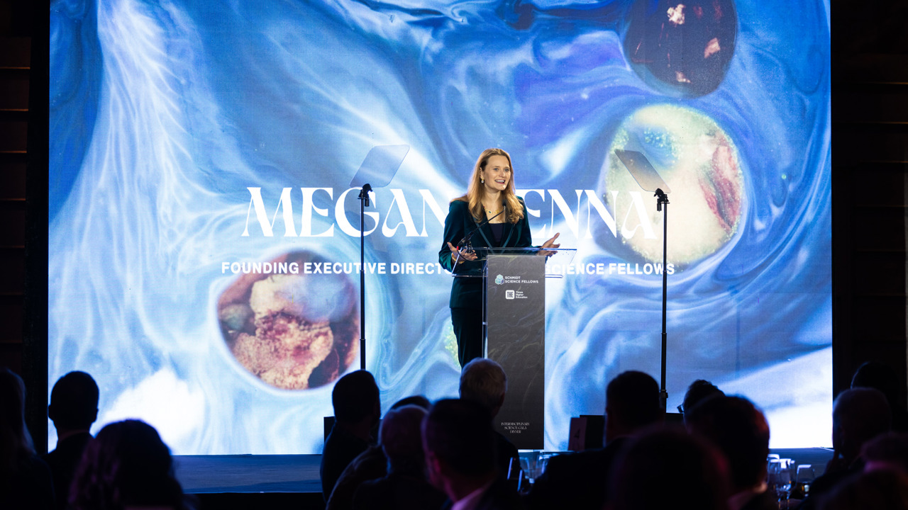 Megan Kenna speaking at a podium in front of a large screen depicting a backdrop of blue swirls and coloured spheres.