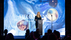 Megan Kenna speaking at a podium in front of a large screen depicting a backdrop of blue swirls and coloured spheres.