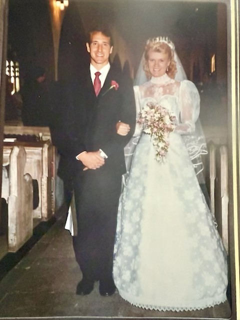 Randy and Rachel pictured standing in the aisle of a church. Randy wears a suit and Rachel is in a white wedding dress holding flowers.
