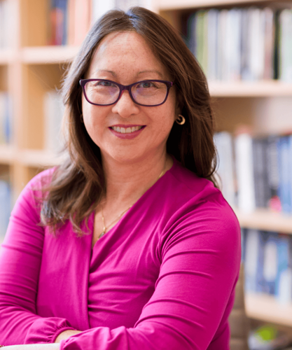 Portrait photo of Karen Yeung, posing in a pink top, and smilign to the camera. Behind her is a bookcase.