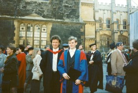Mark in a graduation gown standing next to his supervisor who is also wearing his degree gown. Behind them is the Sheldonian Theater.