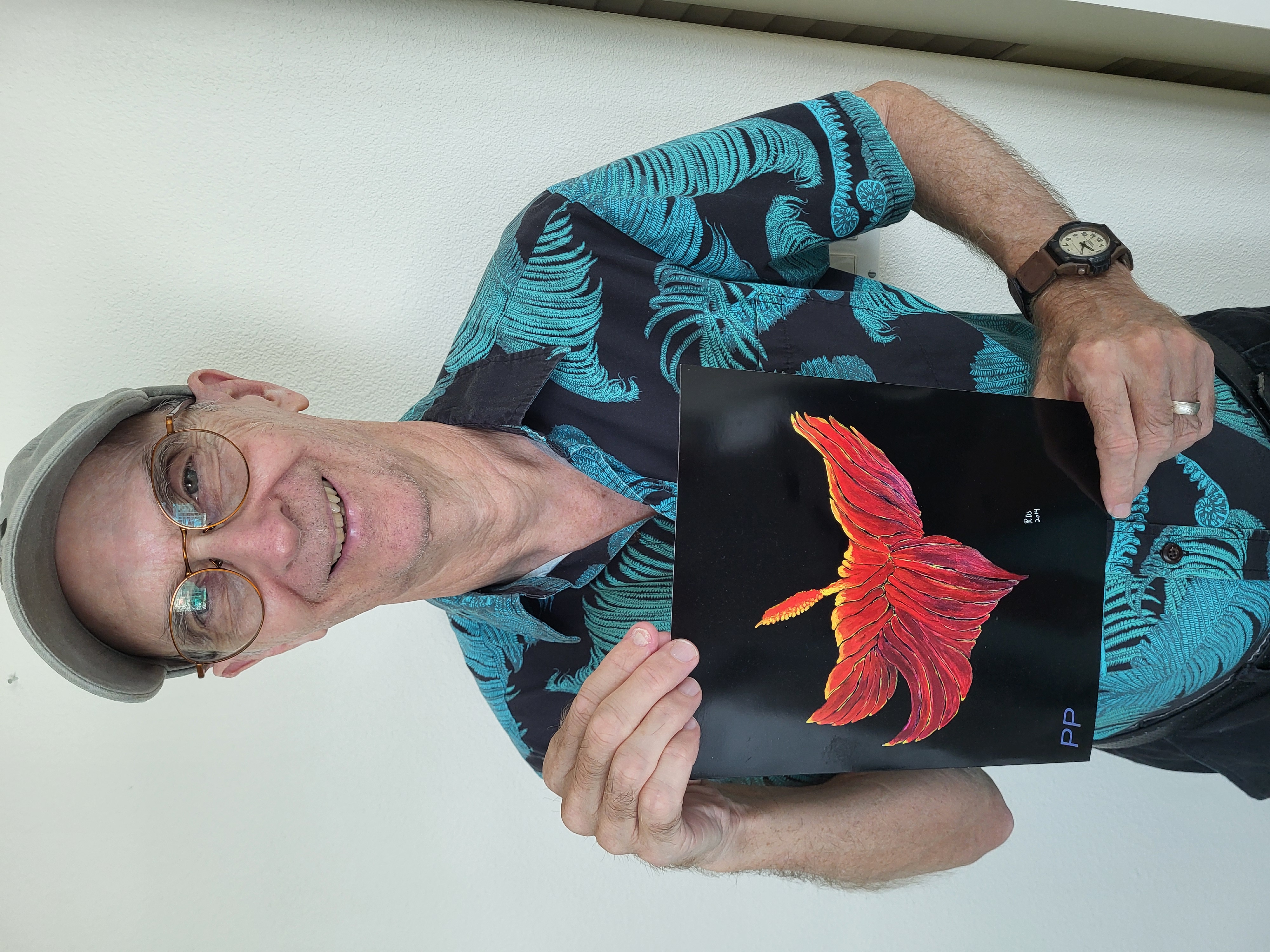 Roger is stood smiling and holding a painting before him. The canvas is black, and a red, orange, and yellow flower has been painted on to it.