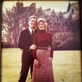 The picture shows Ralph in a black suit standing next to Betsy who is wearing a maroon sweater over a salmon long button-down skirt. They are on a lawn, and there are buildings behind them.