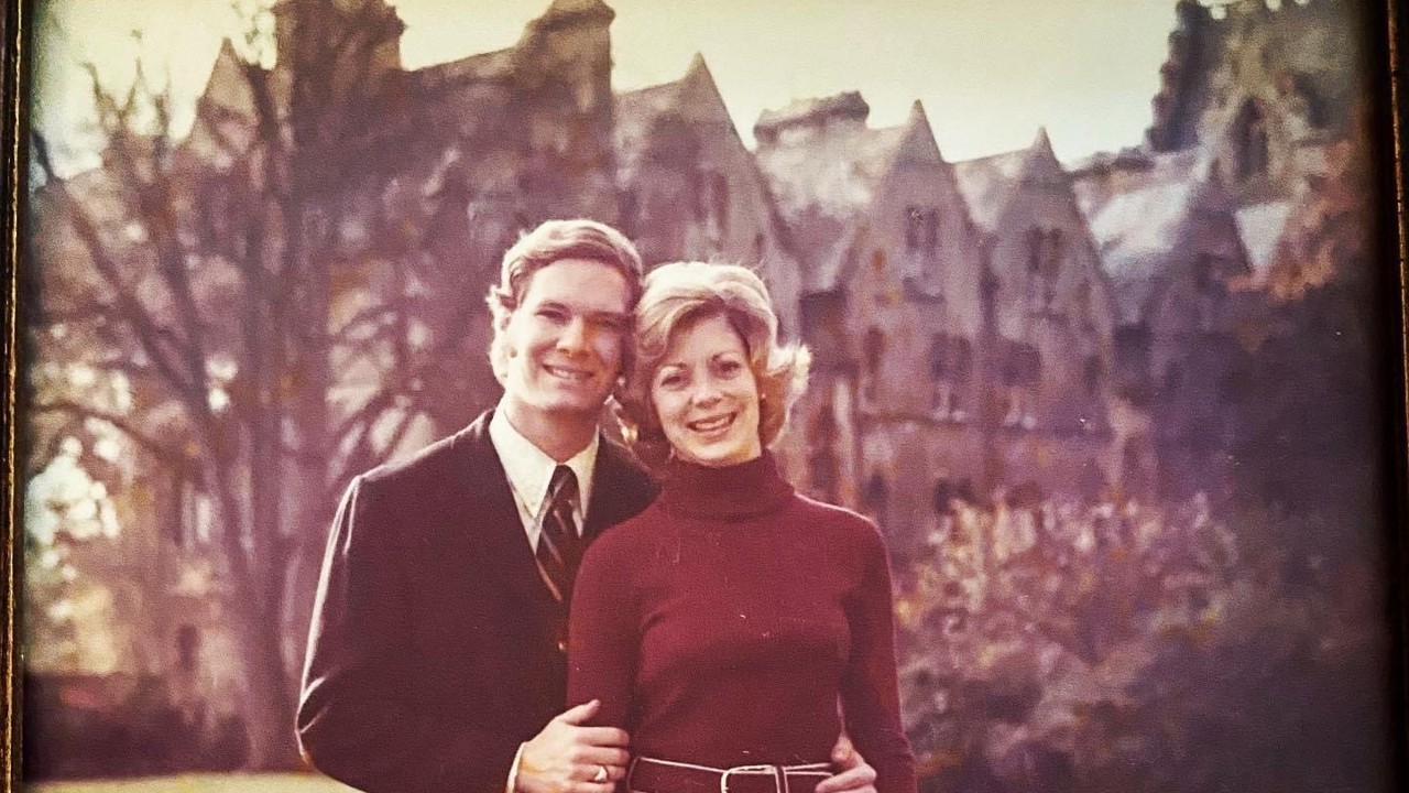 The picture shows Ralph in a black suit standing next to Betsy who is wearing a maroon sweater over a salmon long button-down skirt. They are on a lawn, and there are buildings behind them.
