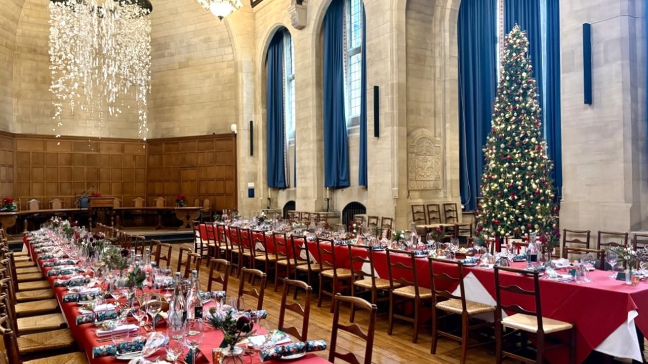 Two long banquet tables in Rhodes House McCall MacBain Hall set for Christmas dinner with a Christmas tree in the bay window