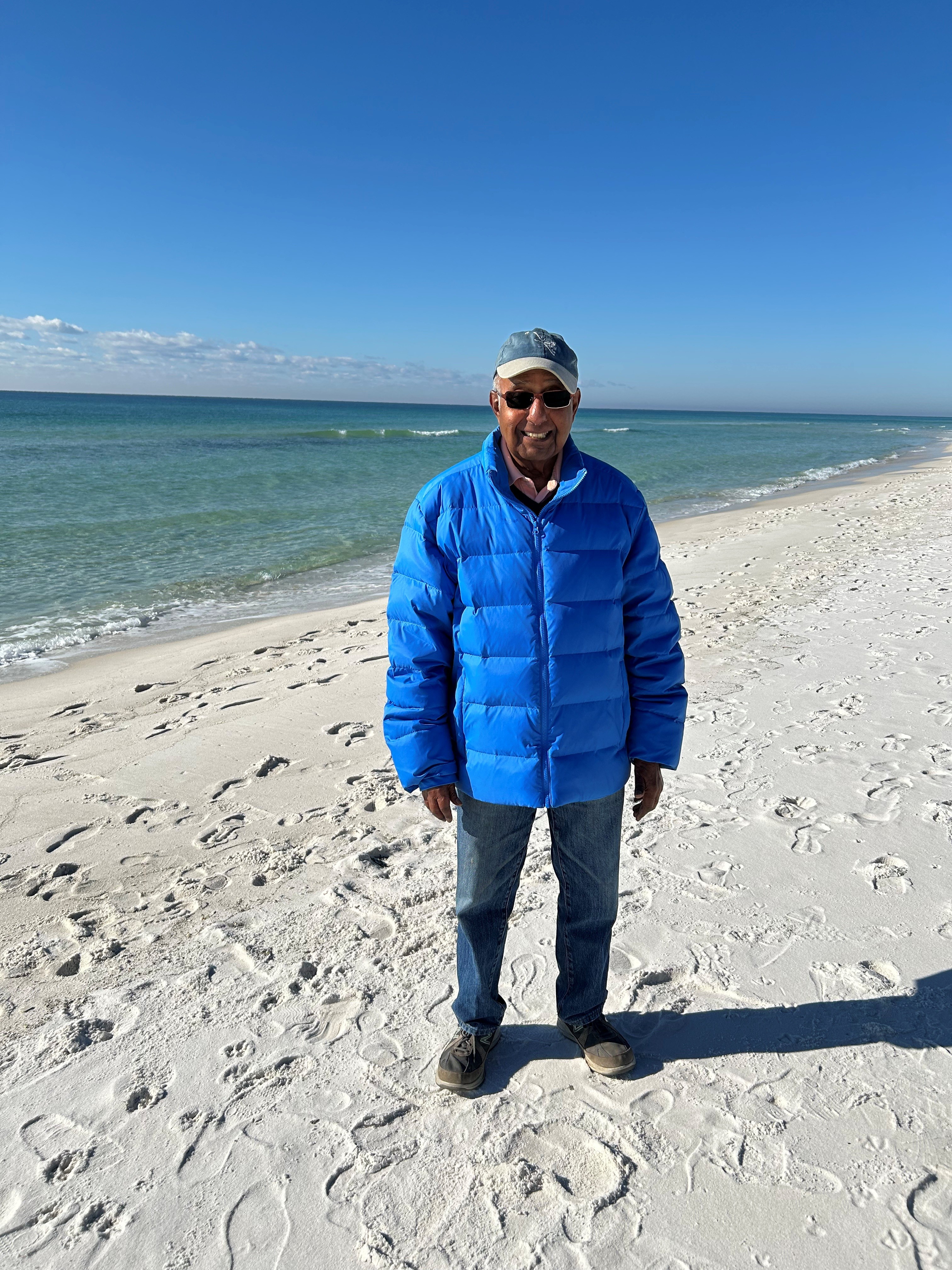 Brij stood on a sandy beach. The ocean can be seen behind him, and he wears a large blue puffer jacket.
