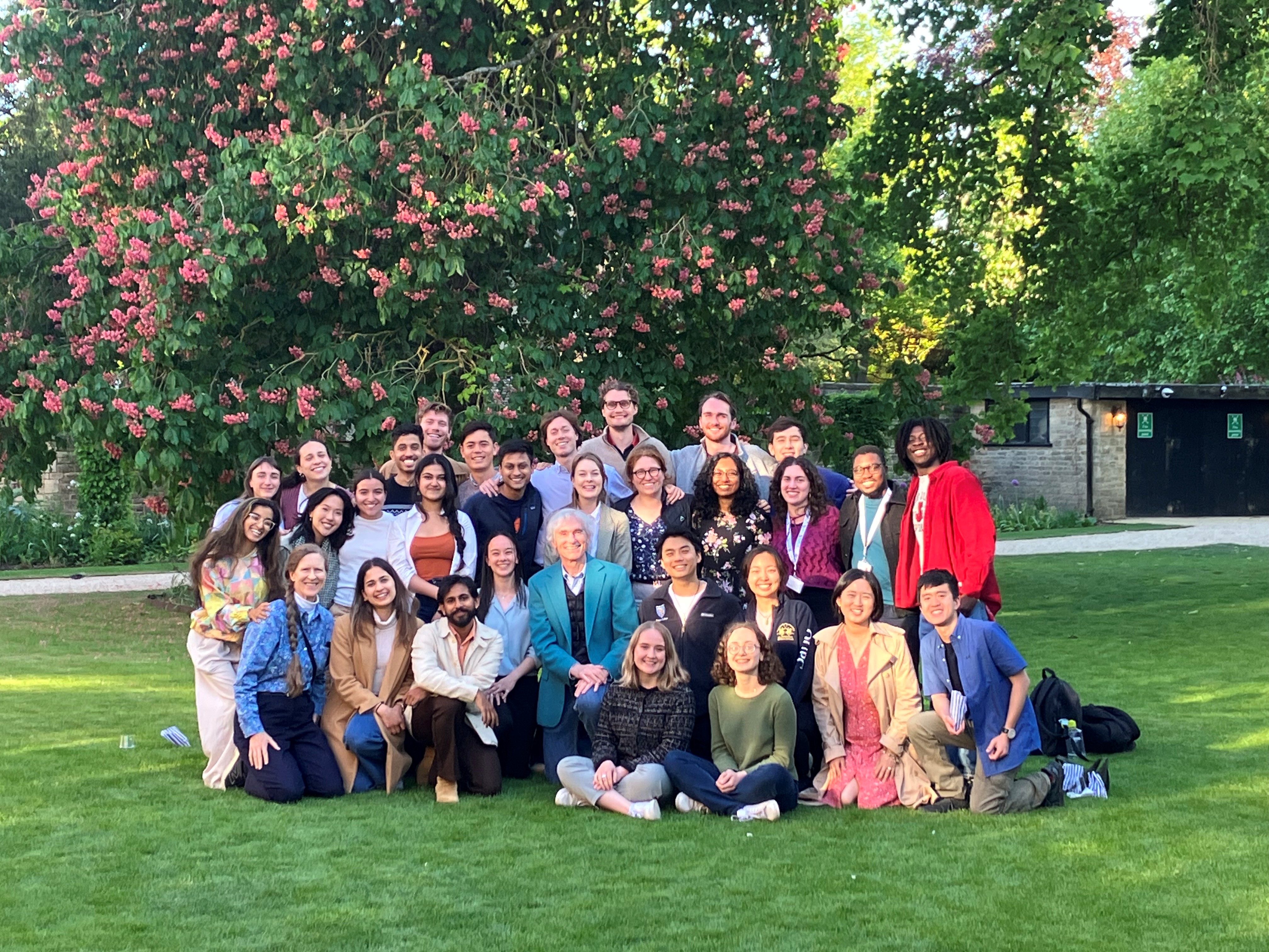 A large group huddled together on a green lawn, with a tree filled wth pink flowers behind them. Tom Barron is in the centre in a blue blazer.