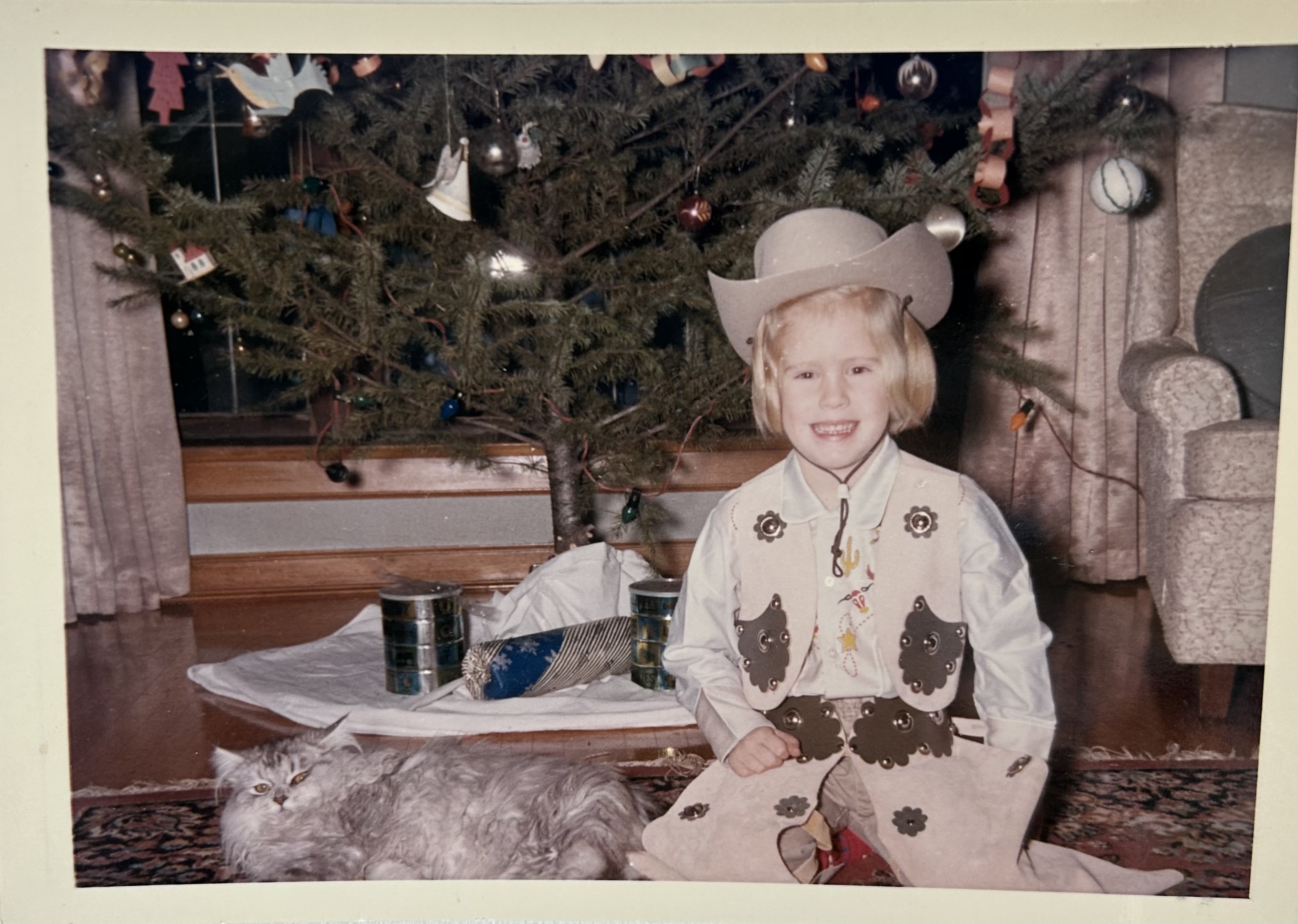 Image of a photograph taken of Meredith as a young girl. She is wearing a cowboy hat, western style waistcoat and trousers. There is a Christmas tree and gray cat to her left.