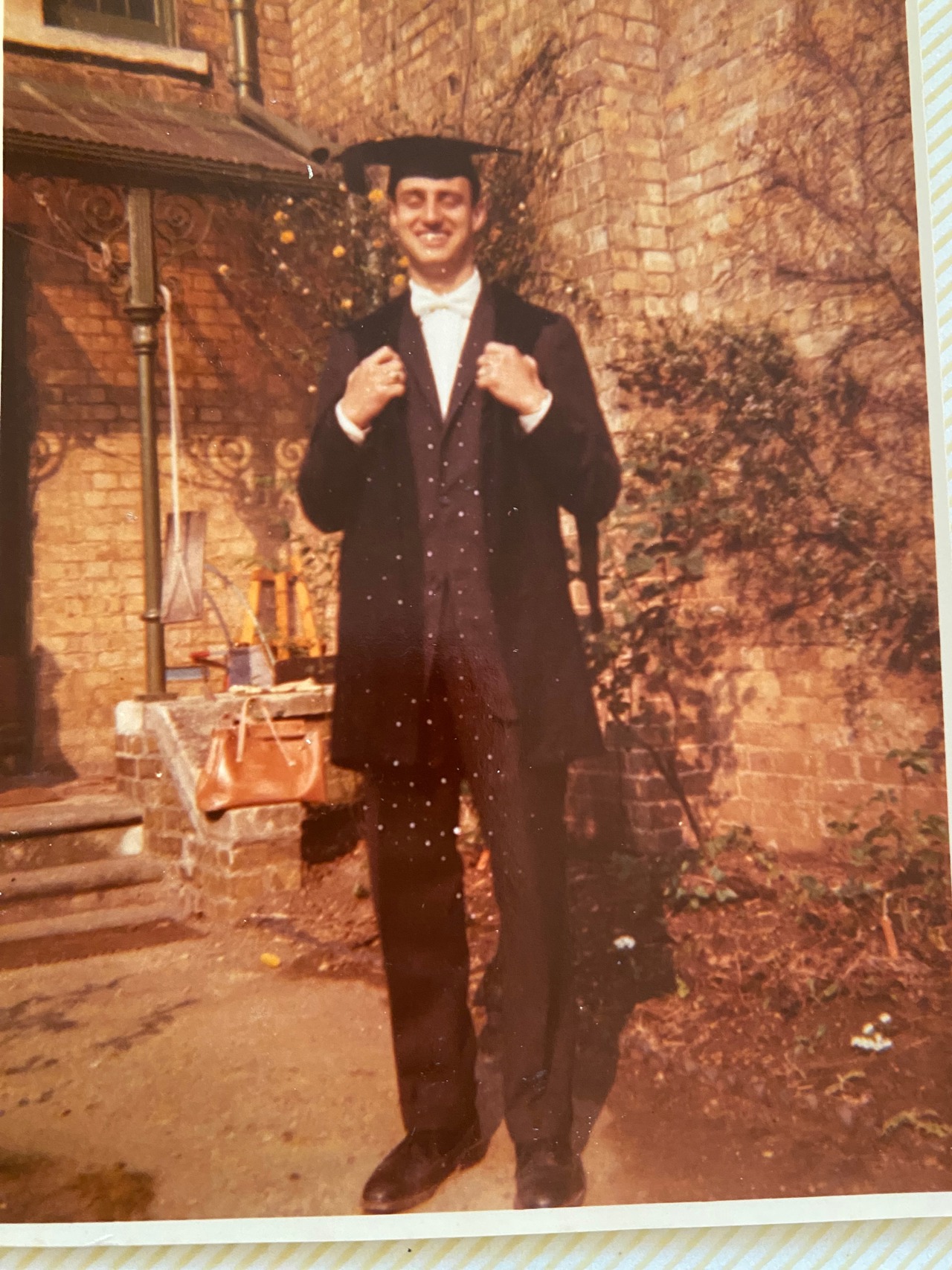 Full length photo of Erich stood smiling wearing his graduation clothes as a young student.