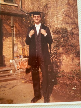 Full length photo of Erich stood smiling wearing his graduation clothes as a young student.