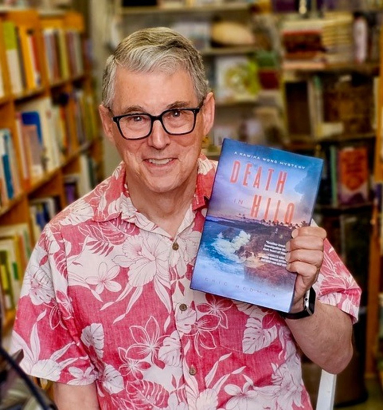 The picture shows Eric in a pink floral shirt holding his book 'Death of Hilo'