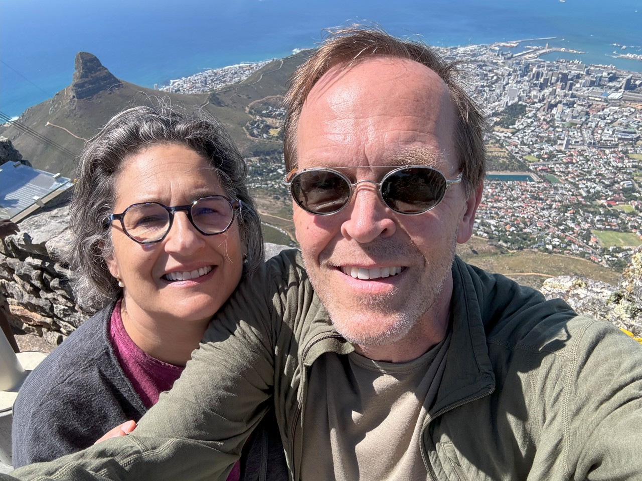 Selfie photo of Eric and his wife. They appear on the top of a mountain as behind them we see a city view, hills, and the ocean. They are both smiling.