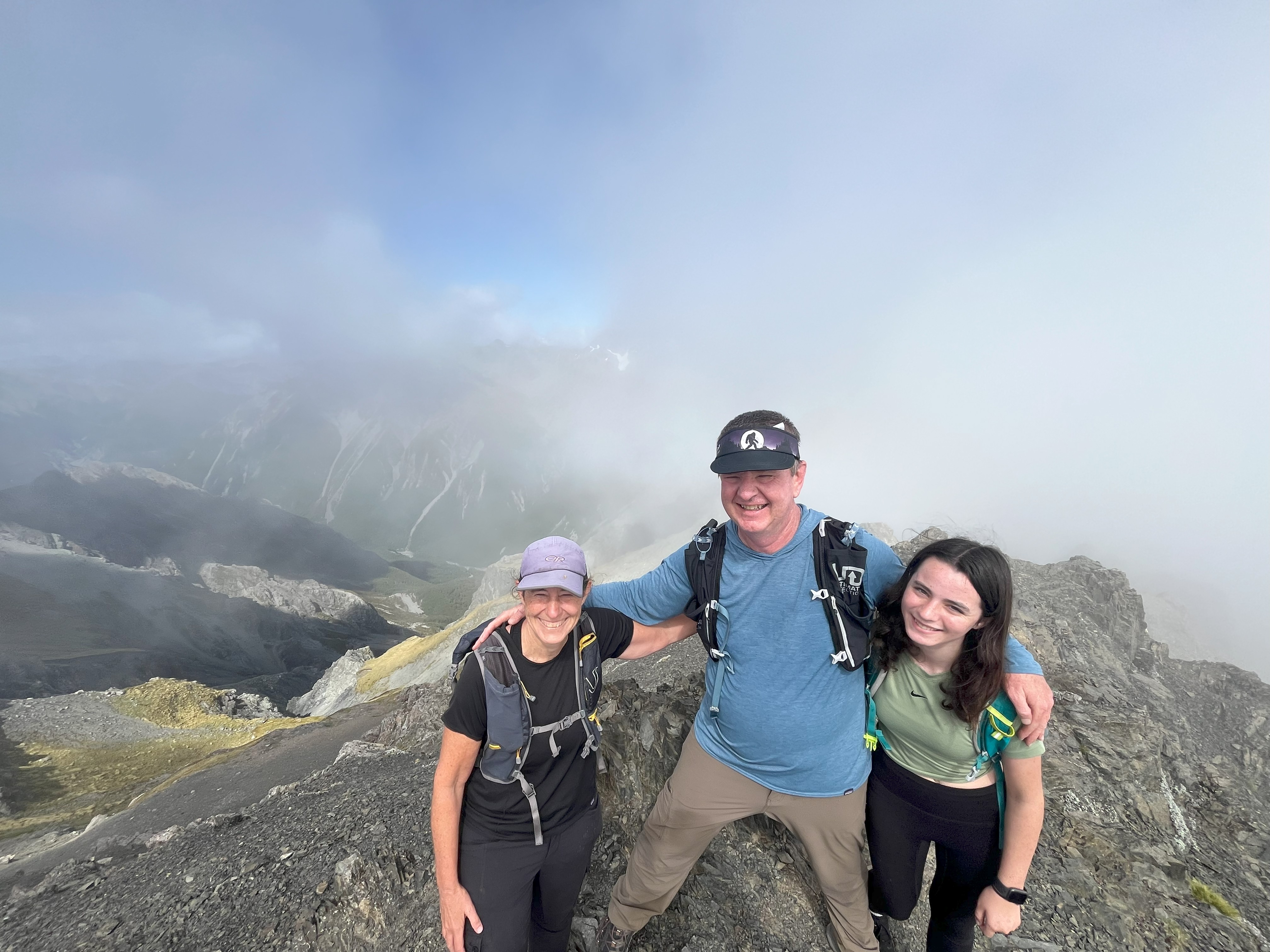 Jane stood to the left wearing a cap and backpack. A man is in the middle and young girl to the right, all have arms around each other. They are at the top of a mountain, you can just about see blue sky and mountain peaks behind them, as there is a lot of mist.