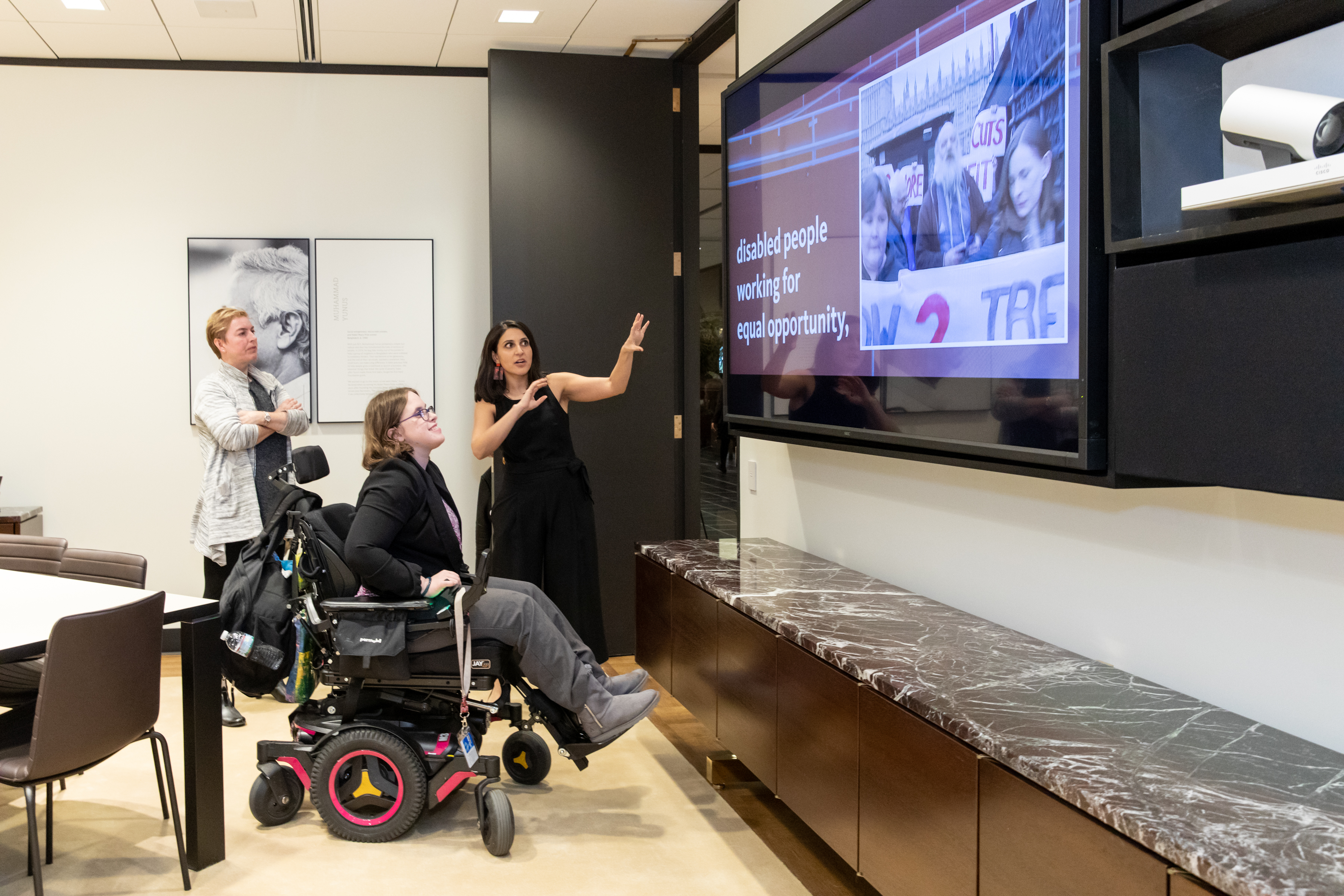 Noorain is seen speaking with a lady in a wheelchair and another lady, who are both looking at the PowerPoint screen Noorain is pointing to which reads 'disabled people working for equal opportunity'