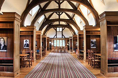 An image of the Rhodes Roseberry Room, which is a long, wooden structure with a vaulted ceiling, a striped carpet down the middle, and wooden sections on both sides with tables and chairs for study.