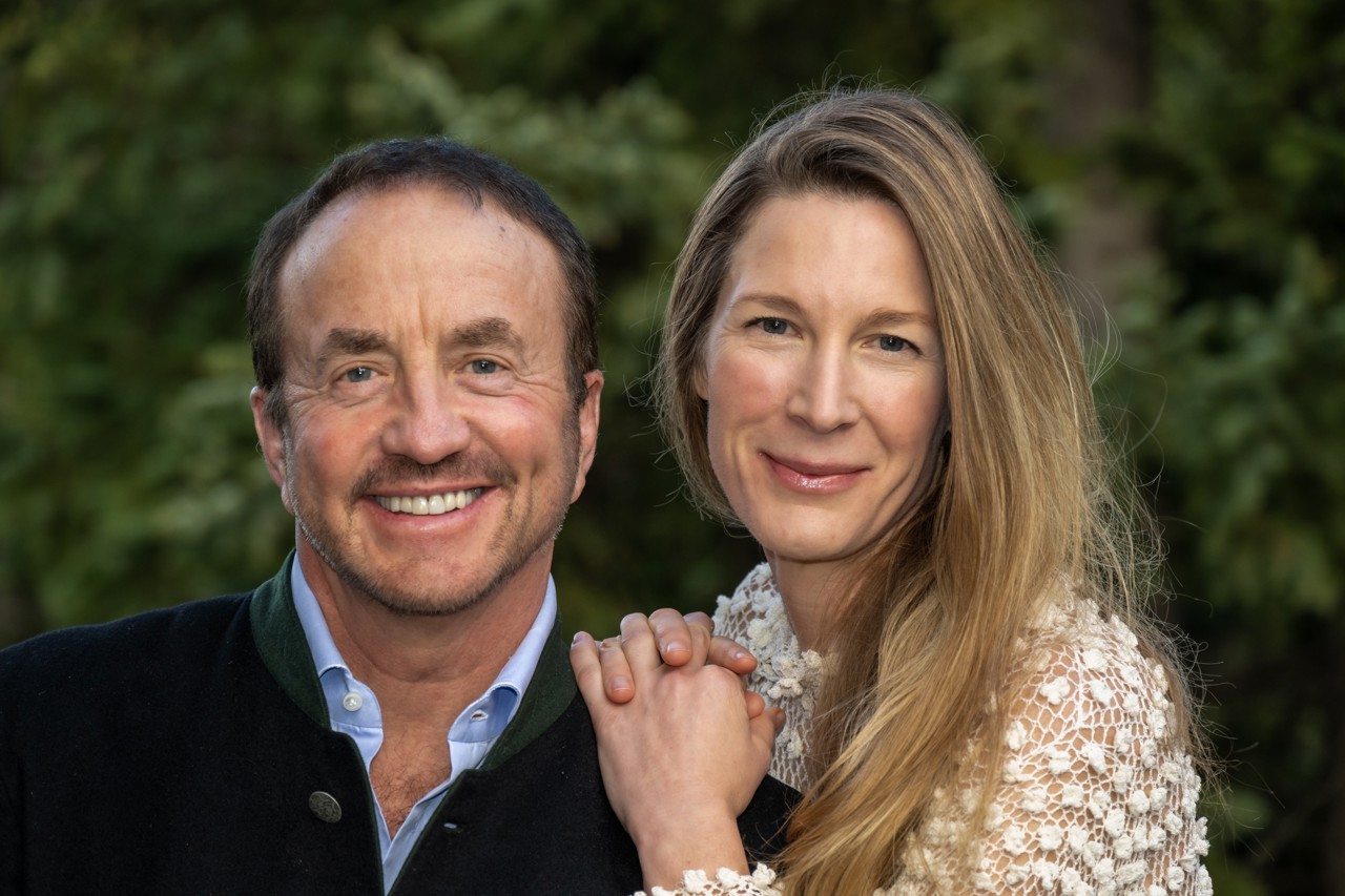 John stood smiling with Marcy holding her hands over his shoulder and looking into the camera.
