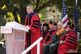 Bob wearing a red gown, stood at a podium, with gowned staff sat behind him before an American flag.