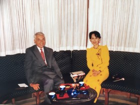 The picture shows Aung San Suu Kui in a yellow dress sitting with Moni in a suit. They are on a couch with a table in front of them.