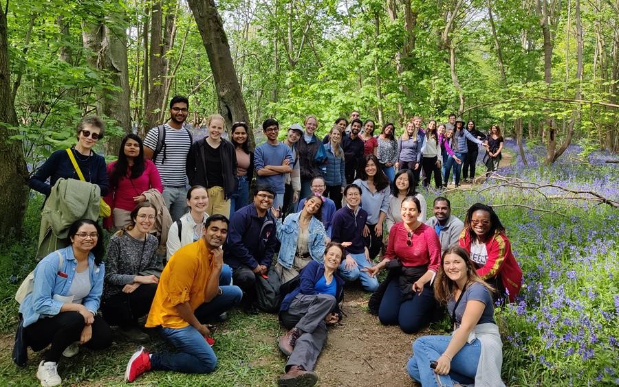 A group of Scholars in the Bluebell woods, surrounded by bluebells. All are smiling and posing for the camera, Elizabeth is laying on the ground, in the centre of the photo, and smiling, with Scholars around her.