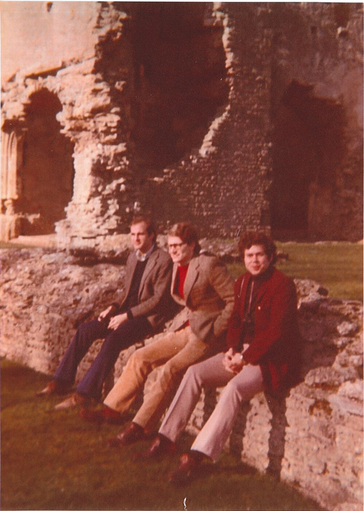 The picture shows a group of three men sitting on a crumbling wall while a huge dilapidated stone building looms in the background