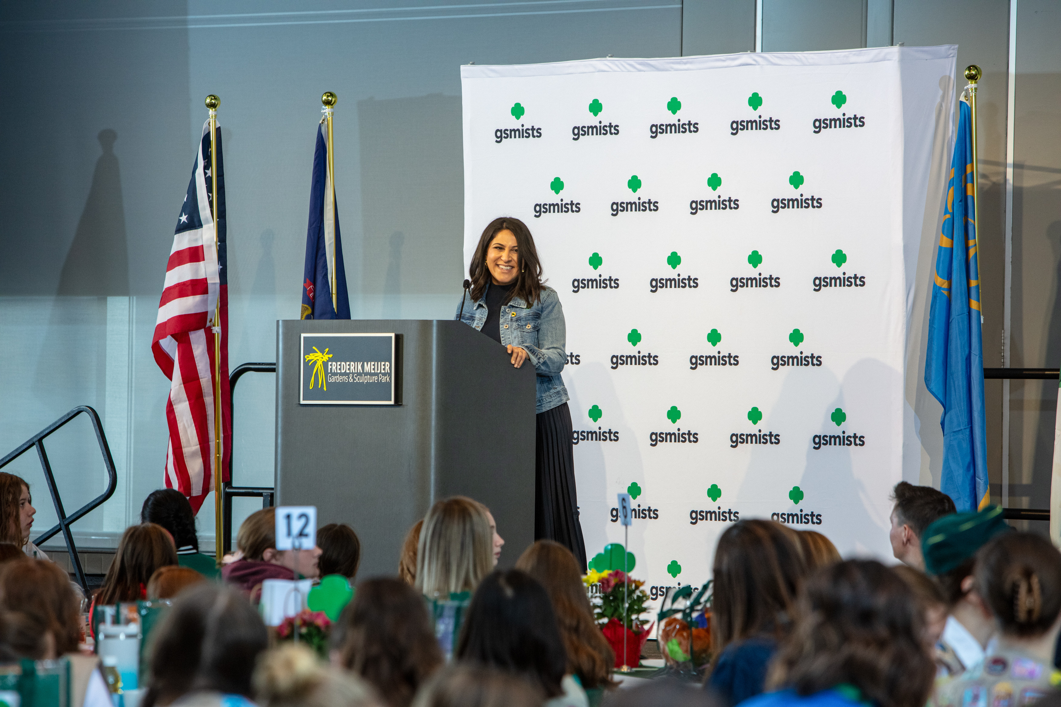 Noorain is stood on a stage before a lecturn and is smiling towards the audience who are sat before her. Behind her is a banner that reads 'gsmists' which is a brand, and there is also the US flag.