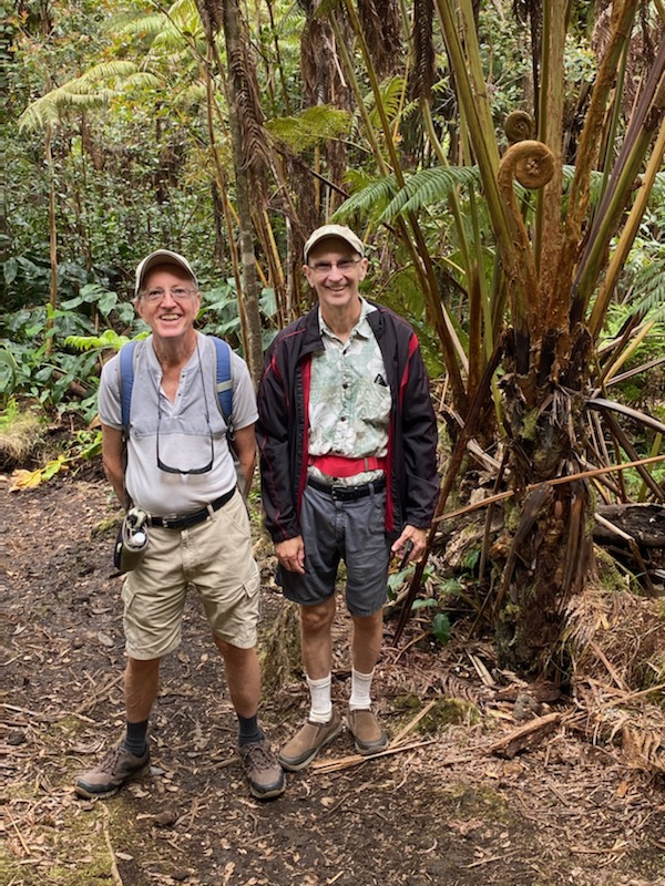 Roger stood to the right of his husband in the rainforest, with trees and greenery behind them. 