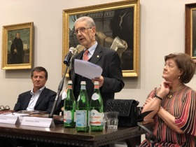 Jaynie sitting at a table while a man in a suit gives a speech. There are several paintings on the wall behind them.