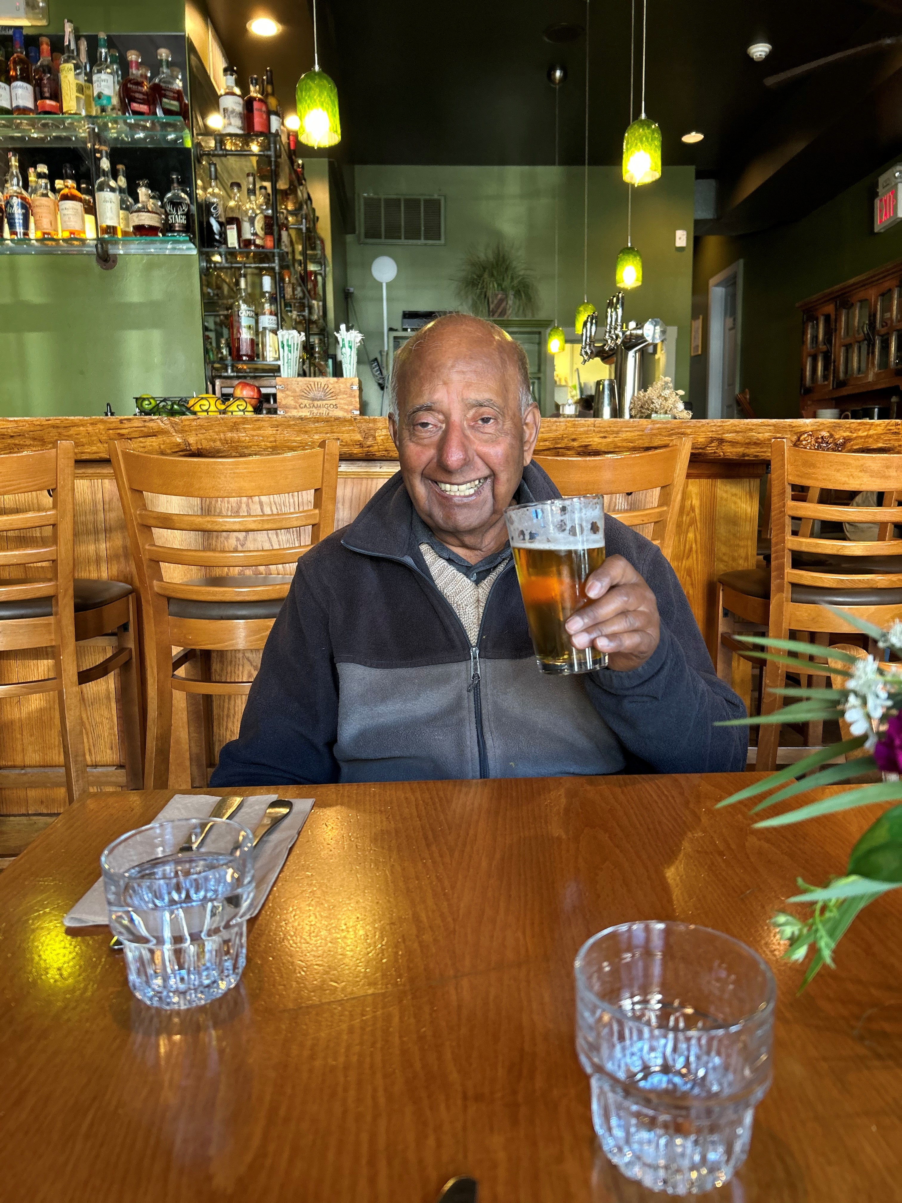 Brij is sat at a table in a restaurant holding a pint of beer and smiling.