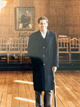 Jonathan stood in a long coat, holding an umbrella, in the hall at Rhodes House. Wooden chairs and portrait behind him.