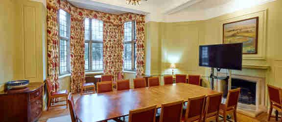 A whole room shot of the Atlantic Room in Rhodes House. In the centre of green-walled room is a long table surrounded by red chairs. At the head of the table in front of a fireplace is a large flat-screen TV. A large bay window framed by floral curtains lets light into the room. A golden chandelier hangs from the centre of the ceiling. 