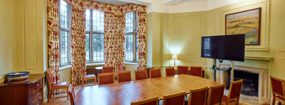 A whole room shot of the Atlantic Room in Rhodes House. In the centre of green-walled room is a long table surrounded by red chairs. At the head of the table in front of a fireplace is a large flat-screen TV. A large bay window framed by floral curtains lets light into the room. A golden chandelier hangs from the centre of the ceiling. 