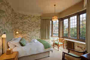 A Bedroom With Green Floral Wallpaper And Green Coloured Soft Furnishings Facing The Bed Are Large Old Fashioned Windows That Allow Light To Flood In (1)